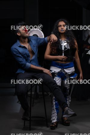 
									Fashion Design, a couple pic , A girl with blue lipstick , dark girl , curly hair , staring , posing , upward eyebrows , straight nose , facial expressions , dark skin , confident , standing , fashionable outfit , wearing a jean jacket , casual outfit , wearing sunglass , smiling , guy siting on a chair , joking