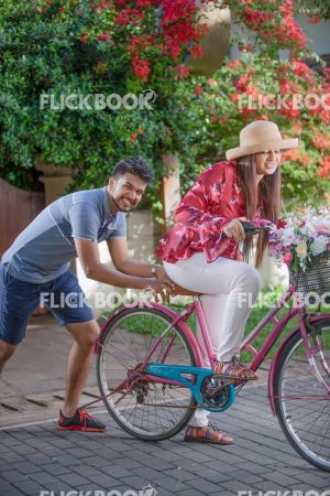 Entertainment, A couple with a flower bicycle , at a garden , wearing a hat , casual outfit , smiling , happy , , pushing the bicycle , trying hard 